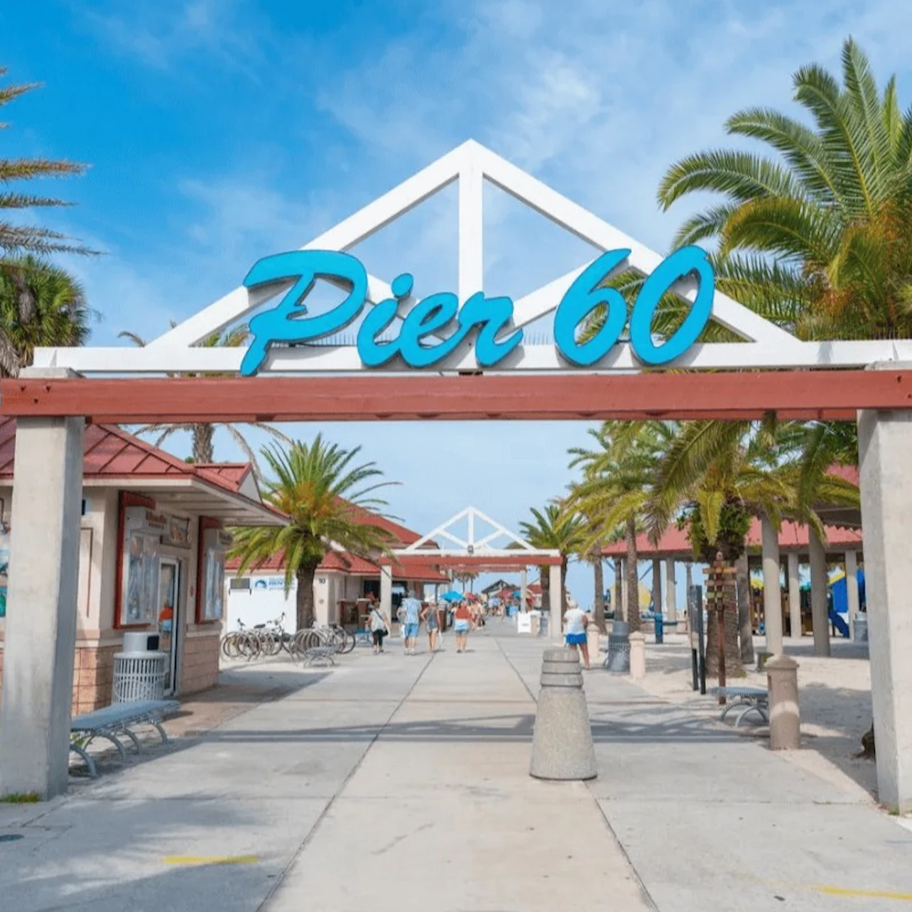 Entrance to Pier 60 with a blue sign, palm trees, and a walkway leading to the beach under a blue sky.