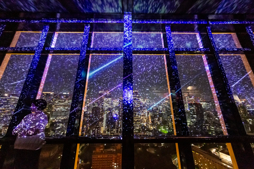 A cityscape at night viewed through large windows with starry lights and beams projected onto the glass.