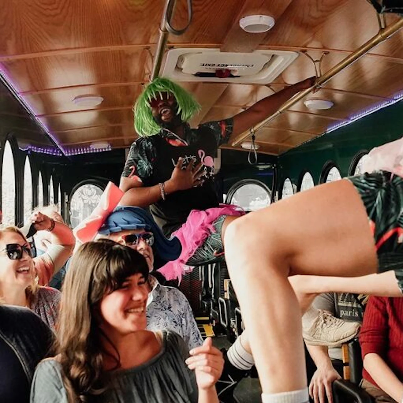 A lively scene inside a bus with people laughing and one individual dressed in a green wig and colorful outfit dancing.