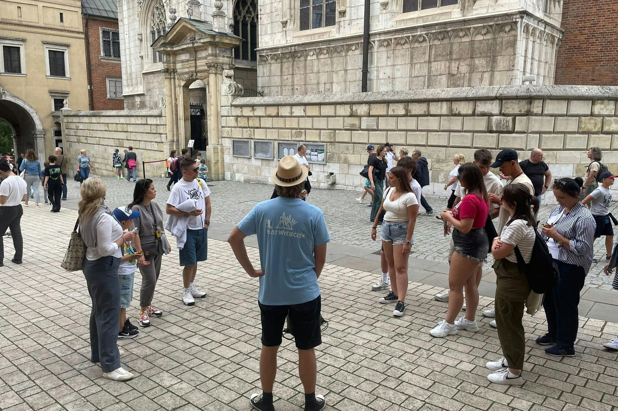 A group of people gathered in a cobblestone square near a historic building, some are listening to a guide wearing a blue shirt.