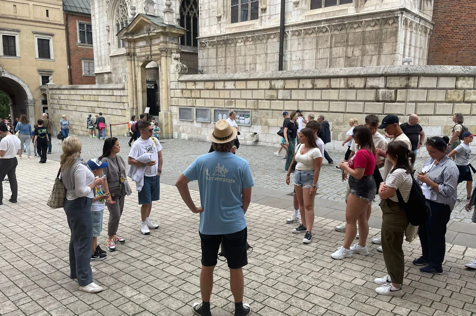 A group of people gathered in a cobblestone square near a historic building, some are listening to a guide wearing a blue shirt.