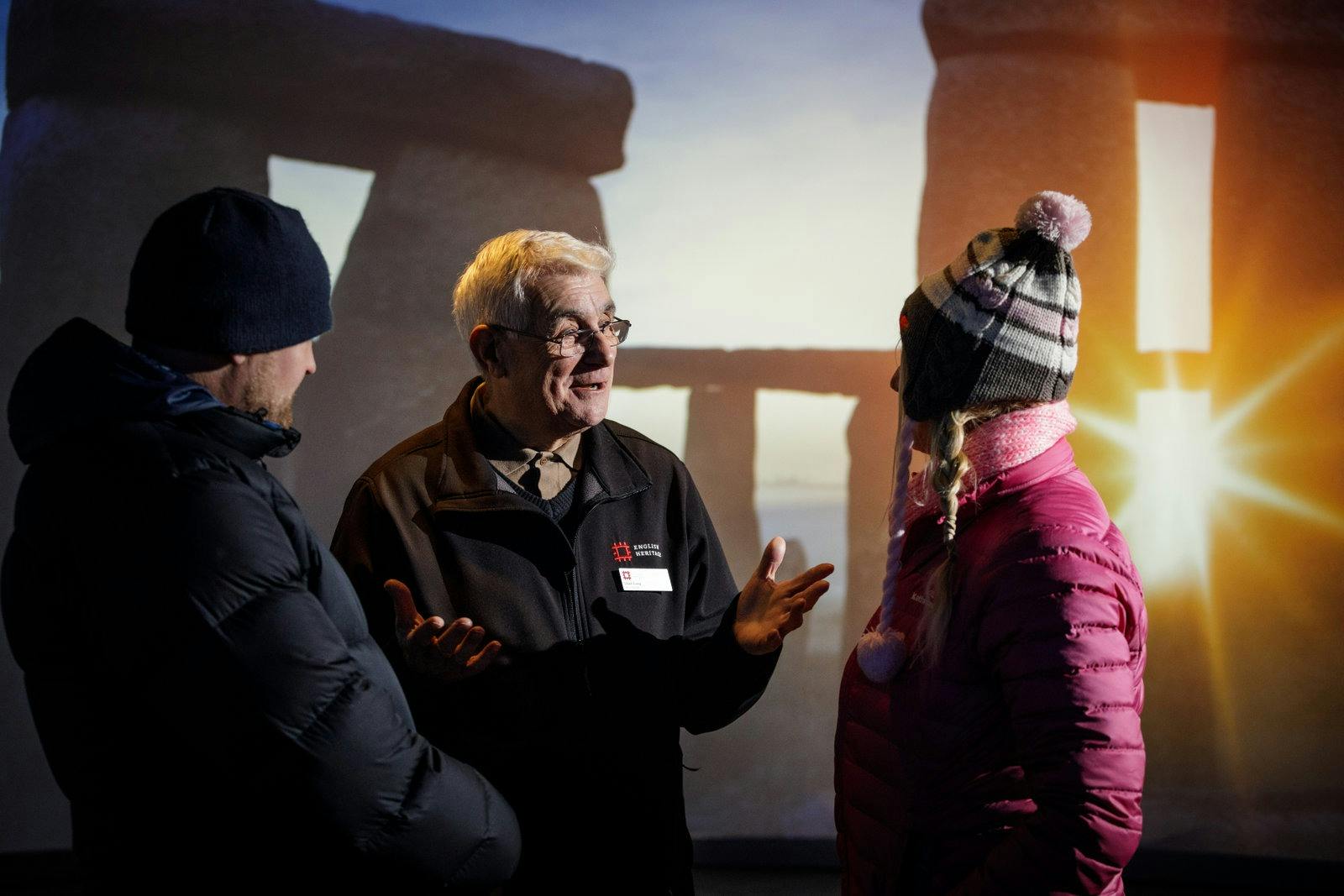 A man wearing a jacket speaks animatedly to a woman and another person in winter attire, with a Stonehenge backdrop.