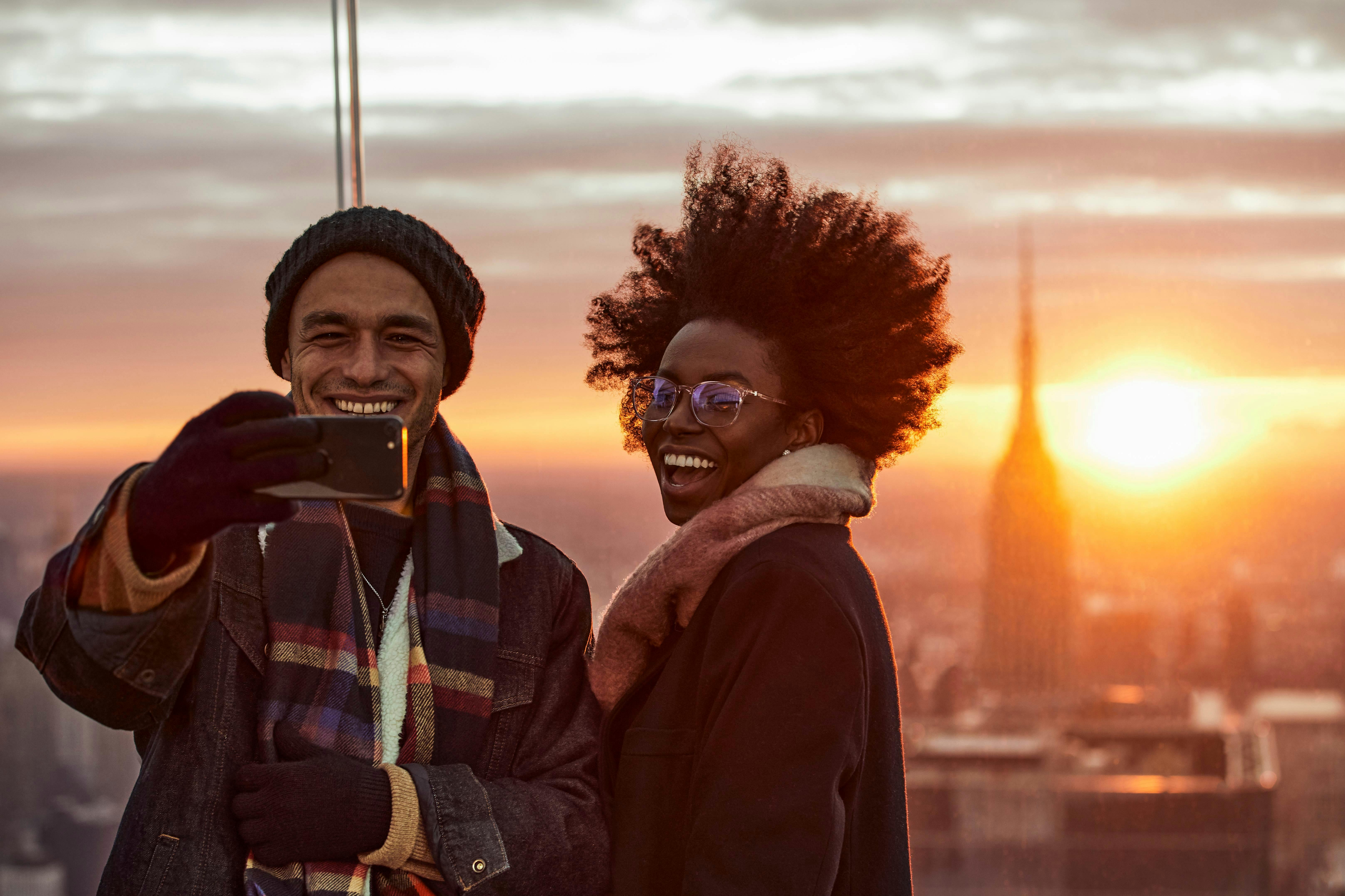 Two people smiling and taking a selfie during sunset with an urban landscape in the background.