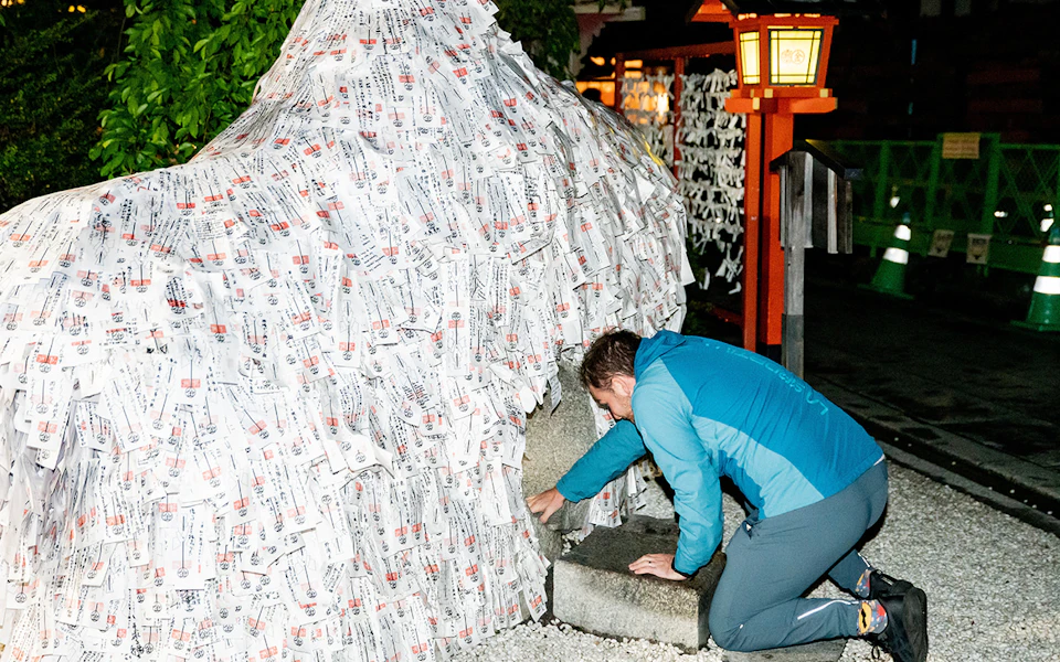 A person in a blue jacket kneeling and crawling through a small stone opening in a structure covered with paper slips at night.