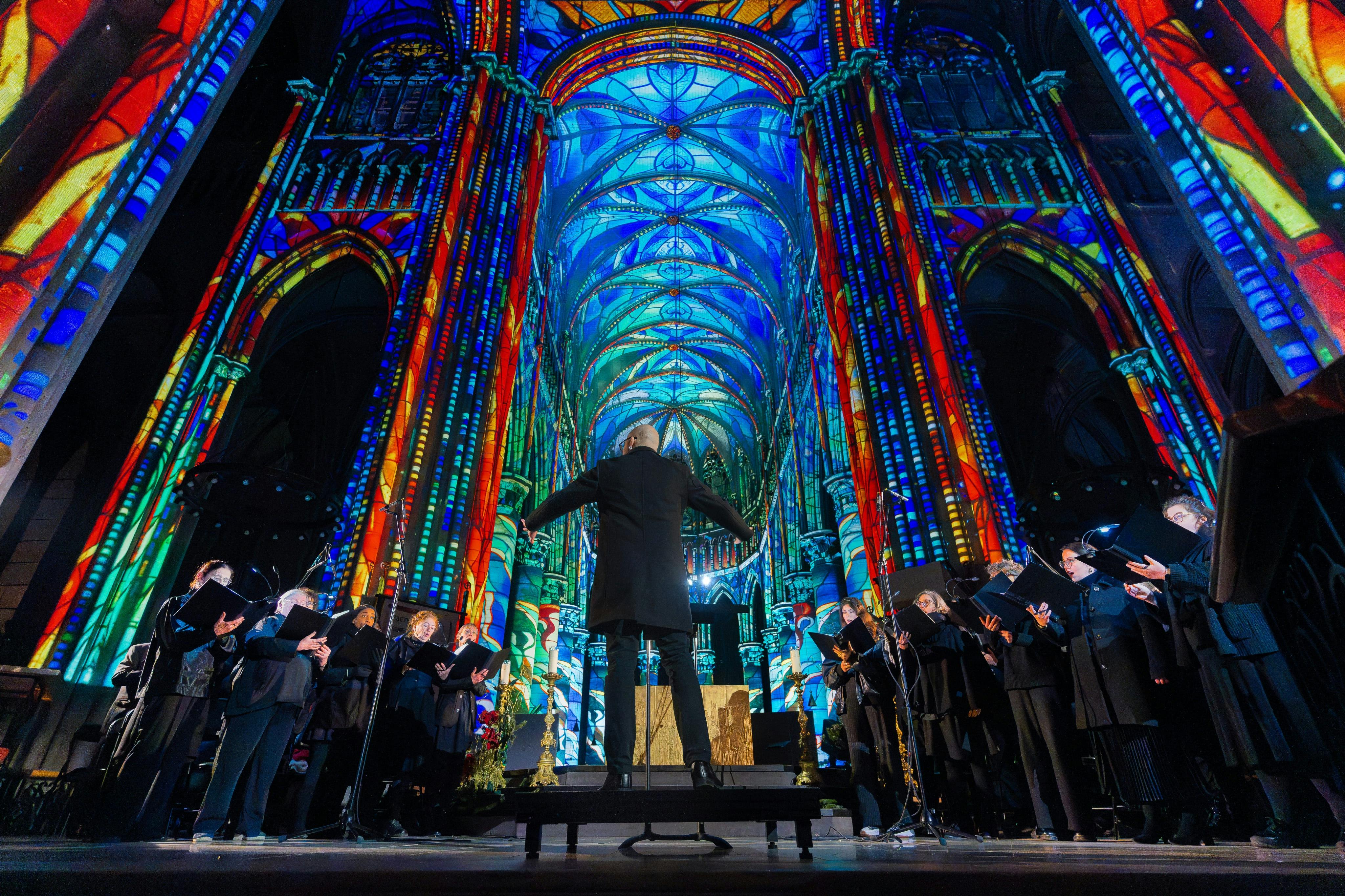 A conductor leads a choir in a cathedral with brightly lit, intricate stained glass patterns projected on the walls.