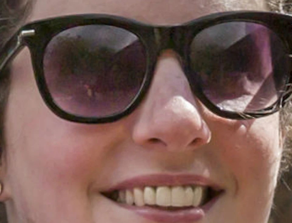 A person with sunglasses smiles while holding a brochure on a double-decker bus near a historic, gothic-style building.