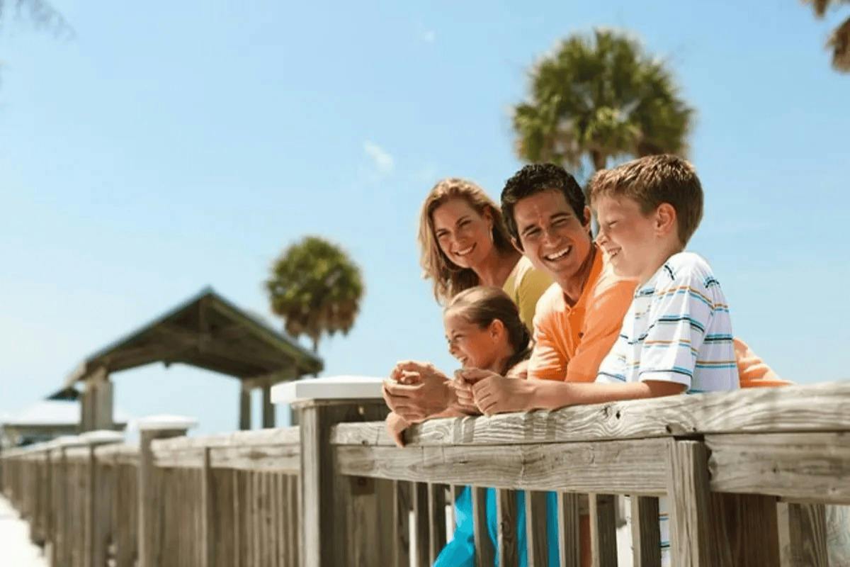 A family of four smiles while leaning on a wooden railing outdoors under a blue sky, with palm trees in the background.