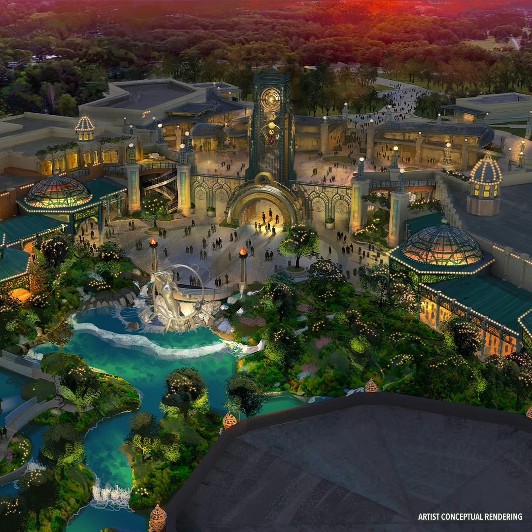 Vista aerea dell'ingresso di un parco a tema al crepuscolo con la torre dell'orologio centrale, gli edifici circostanti, il verde e un piccolo stagno.