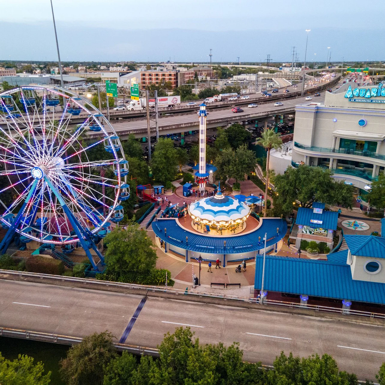 Aerial view of an amusement park featuring a Ferris wheel, a carousel, and a tall ride, with buildings and roads in the background.