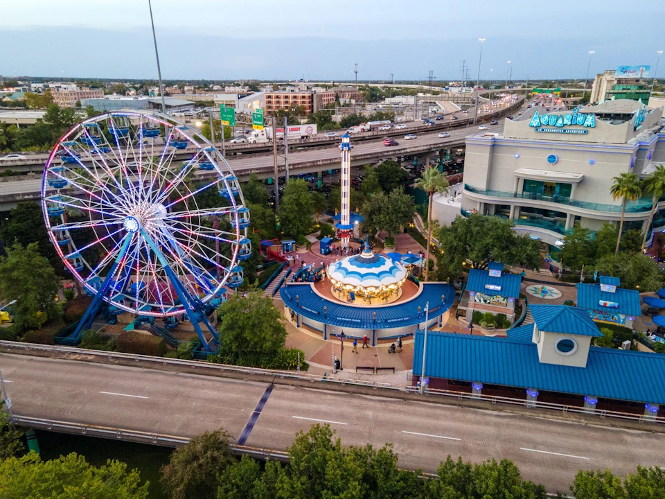 Aerial view of an amusement park featuring a Ferris wheel, a carousel, and a tall ride, with buildings and roads in the background.