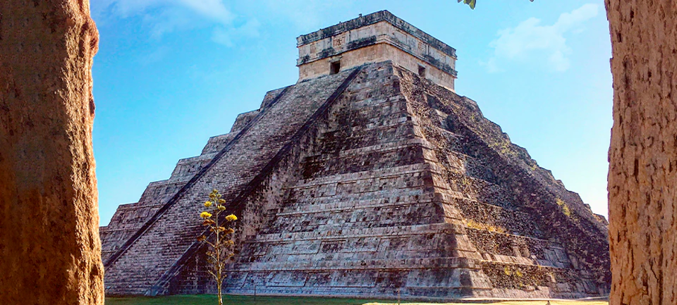 A large ancient stone pyramid with a steep staircase ascends to a small rectangular structure at the top under a clear blue sky.