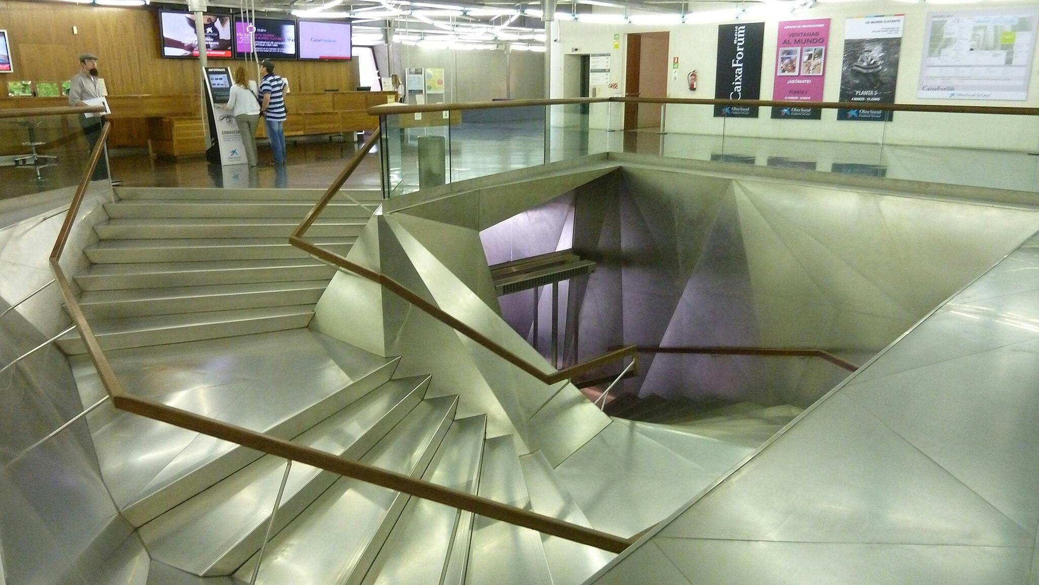 Modern interior with metal stairs leading downward, glass and wood railings, two people near a kiosk, and various wall posters.