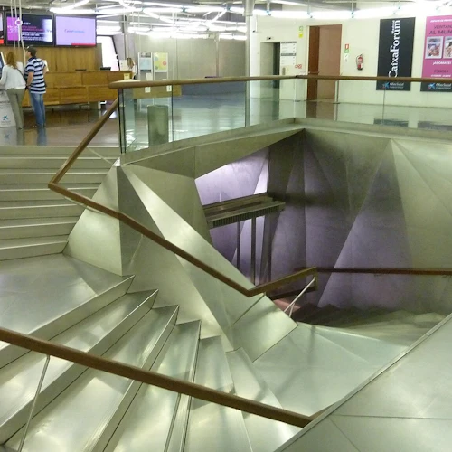 Modern inredning med metalltrappor som leder nedåt, glas- och träräcken, två personer nära en kiosk och olika väggposters.