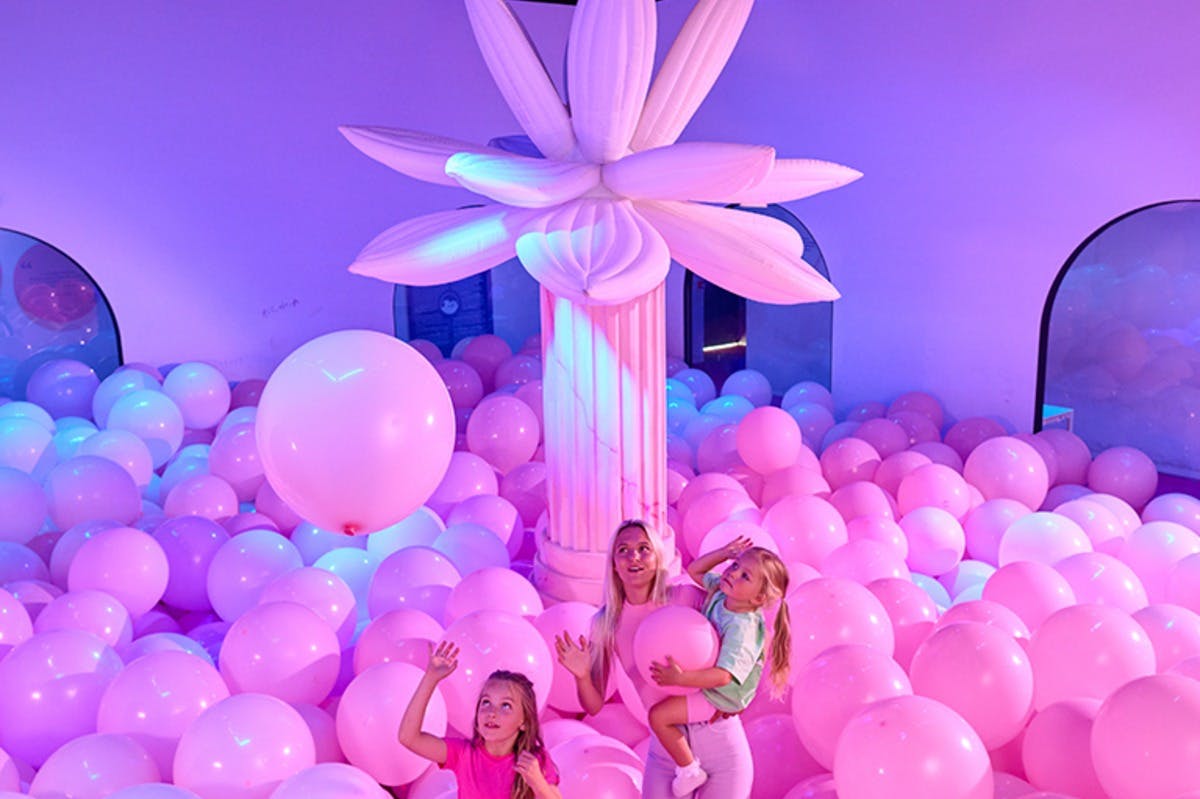 ピンクと紫の風船でいっぱいの部屋で、女性と2人の子供が遊んでいます。隣には、柱に取り付けられた膨張可能な大きな花があります。