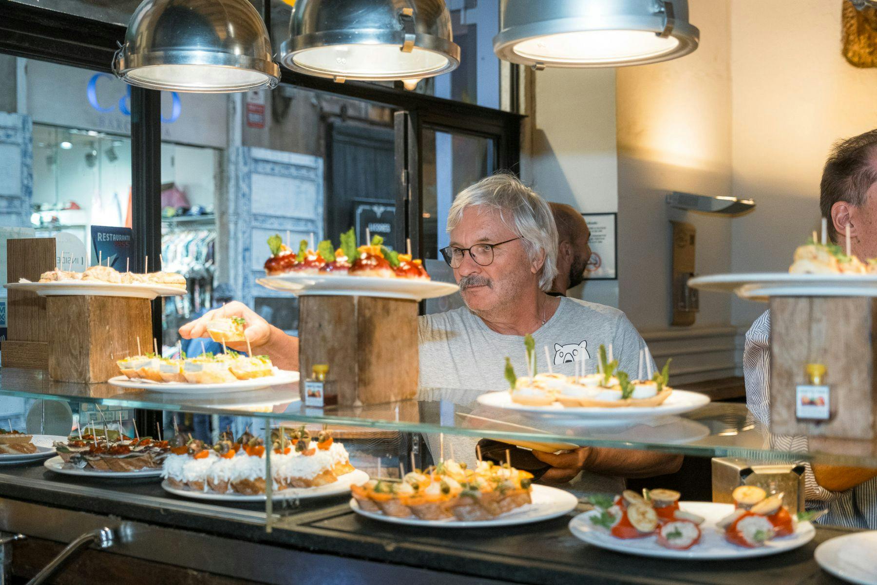 Um homem a selecionar comida de um expositor de pequenos pratos num restaurante bem iluminado, com vários pratos de aperitivos no balcão.