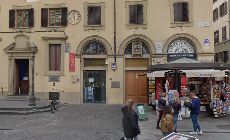 Street view of a building with arched doorways and a museum sign, people walking, and a newsstand on the right.