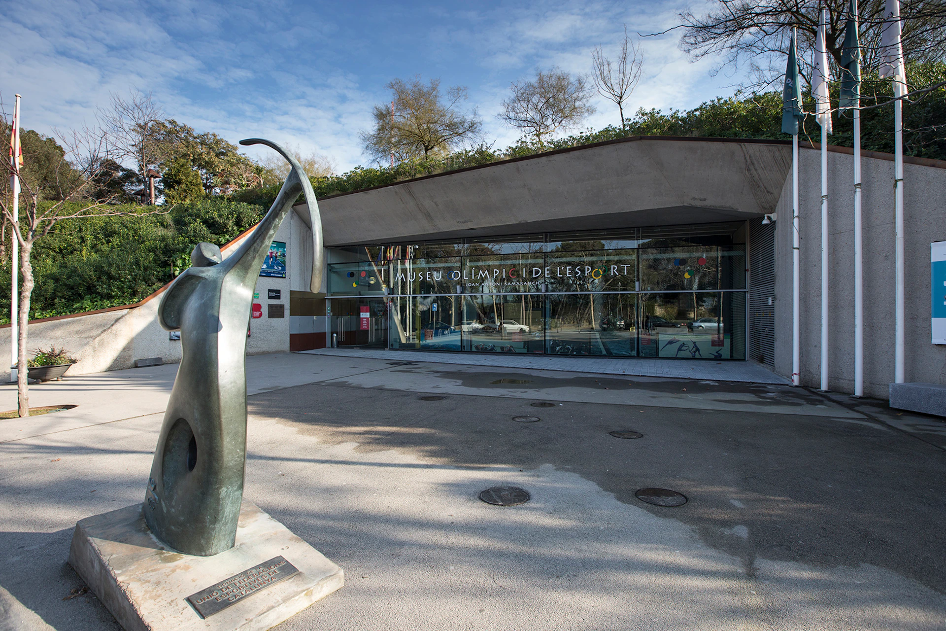 A modern building with glass doors labeled "Museu Olímpic de l'Esport," with a large sculpture of a figure holding a ribbon outside.
