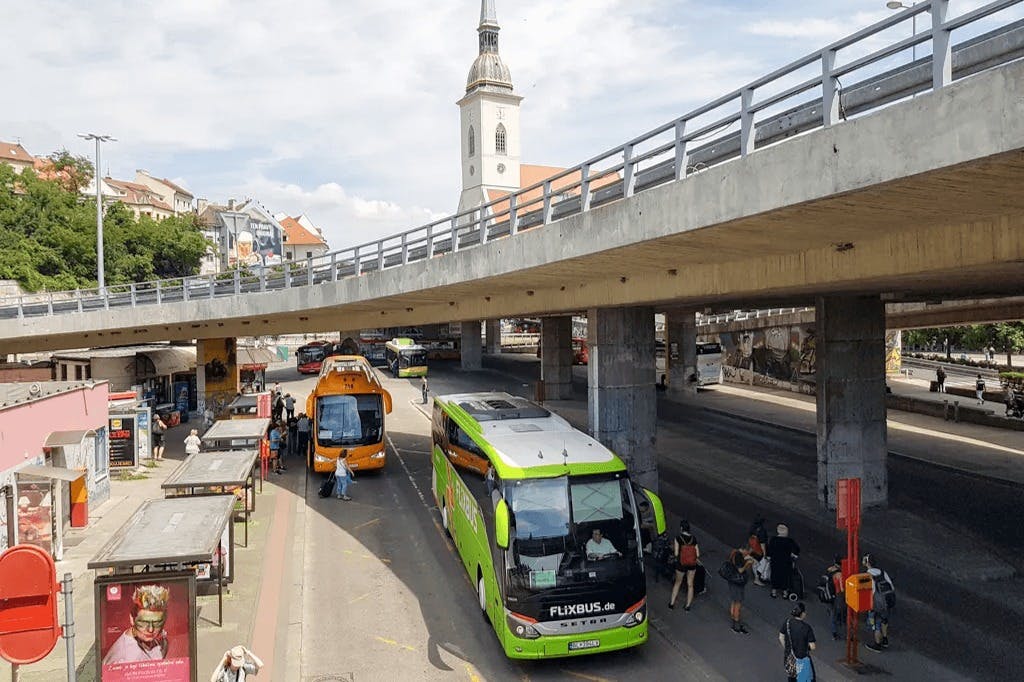 Αρκετά λεωφορεία είναι σταθμευμένα κάτω και κοντά σε έναν υπερυψωμένο αυτοκινητόδρομο από σκυρόδεμα. Στο βάθος διακρίνεται ένας λευκός πύργος εκκλησίας.