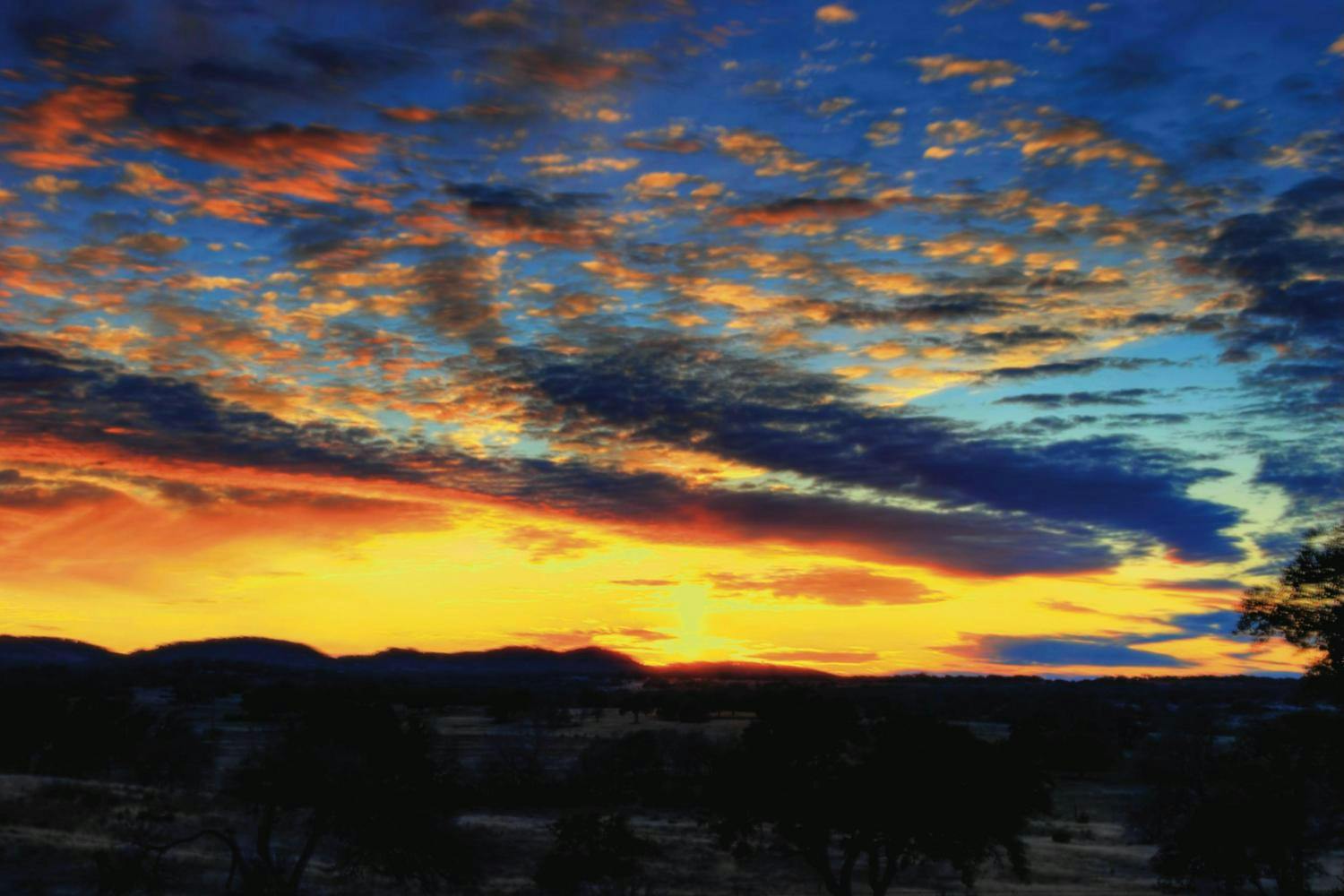 Vivid sunset with a sky full of pink, orange, and blue clouds, silhouetted trees, and distant hills.