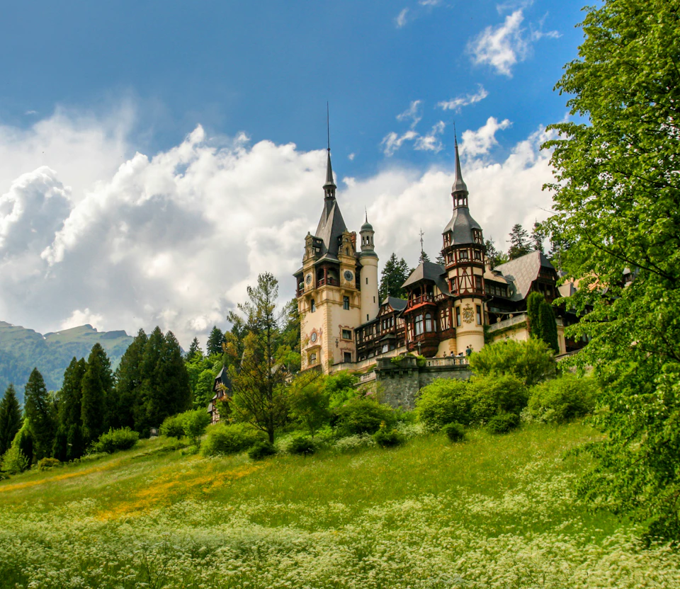 A historic castle with intricate architecture, surrounded by lush greenery and under a partly cloudy sky.