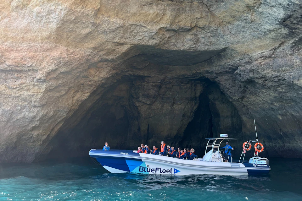 A boat labeled 'BlueFleet' with passengers wearing life jackets is near a rocky cave entrance over clear water.