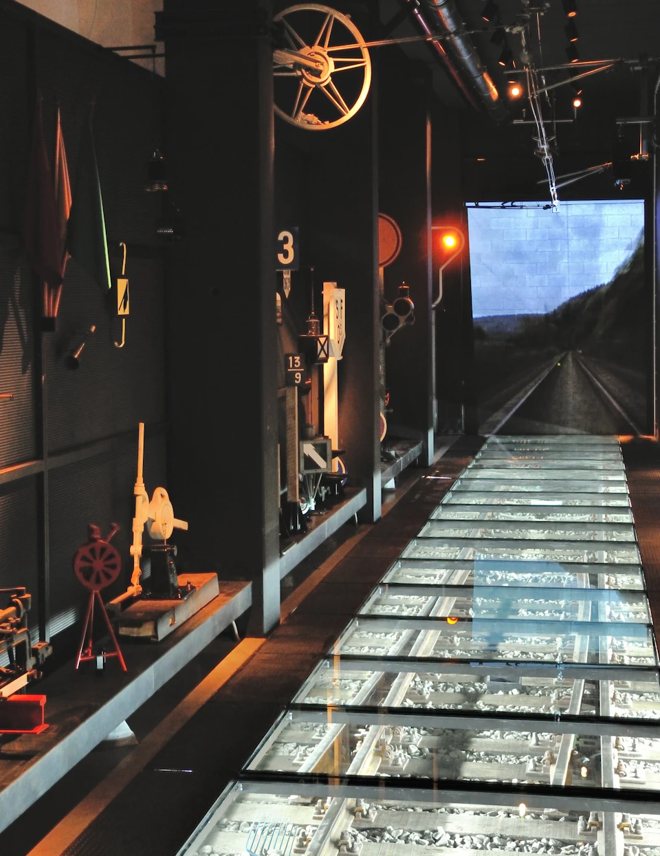 A museum exhibit featuring vintage railroad equipment and a glass floor revealing train tracks, leading to a video screen of a railroad.
