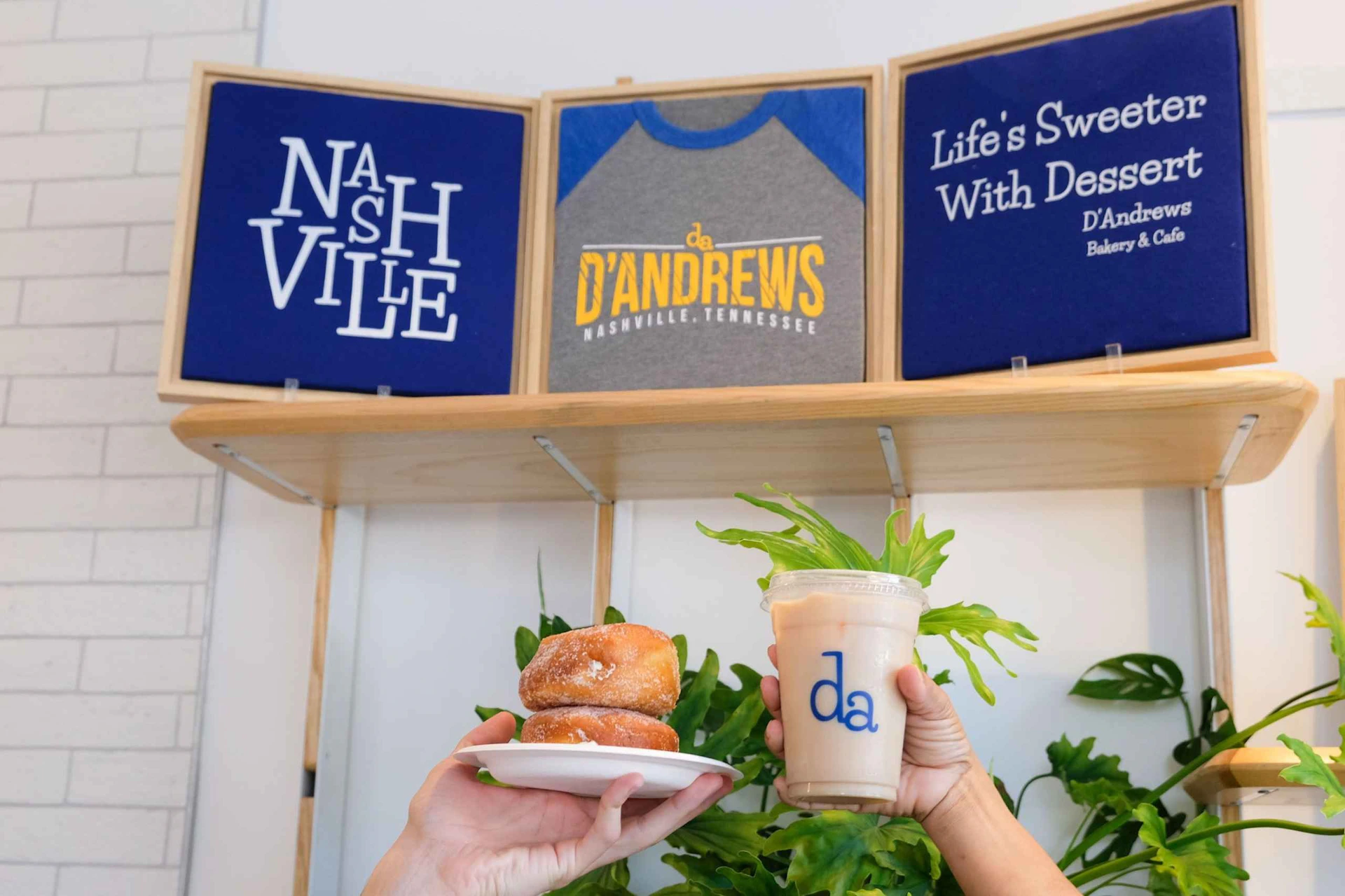 Two hands holding donuts on a plate and an iced drink in front of a shelf with three signs displaying Nashville-related text.