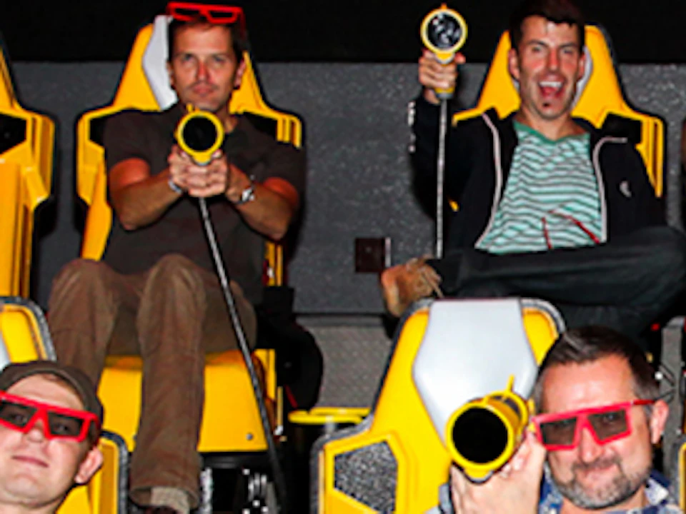People wearing 3D glasses seated in yellow motion simulator chairs, holding light guns, smiling and engaged in an interactive ride.