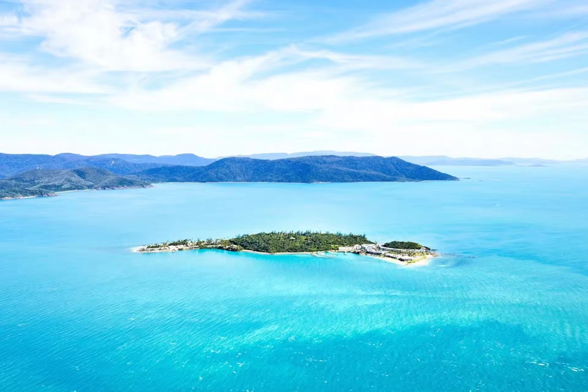 Aerial view of a small, lush green island surrounded by turquoise water, with a larger landmass visible in the background.