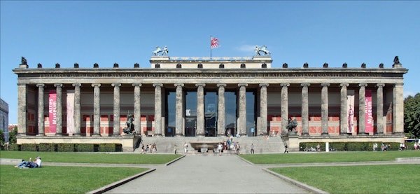 A neoclassical building with tall columns, sculptures, and a red flag, surrounded by visitors on a sunny day.