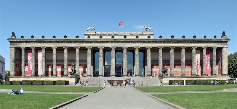 A neoclassical building with tall columns, sculptures, and a red flag, surrounded by visitors on a sunny day.