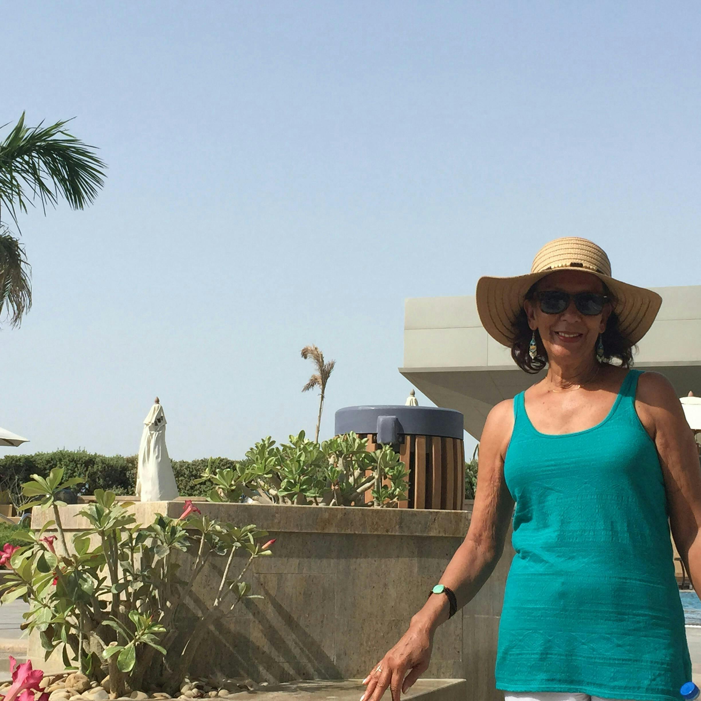 A smiling woman in a turquoise tank top and wide-brimmed hat stands outdoors near plants, with a building and palm tree in the background.