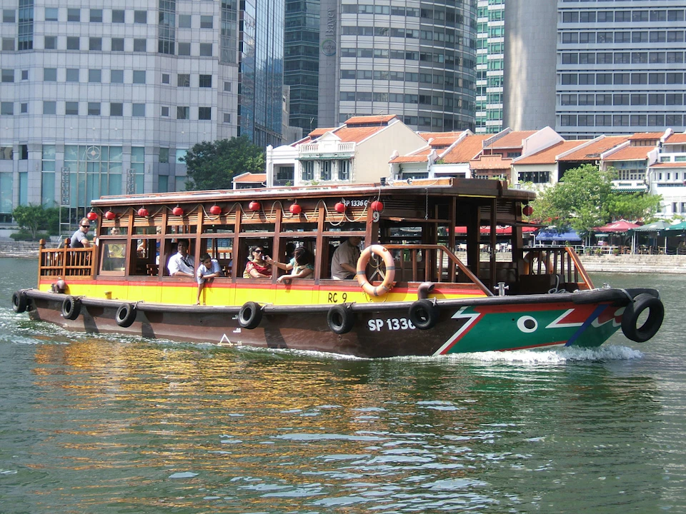 A wooden passenger boat with people aboard travels on a river in front of modern buildings and traditional houses.