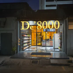 A storefront at night with the illuminated sign "De'8000." Shelves of items are visible through the glass doors.