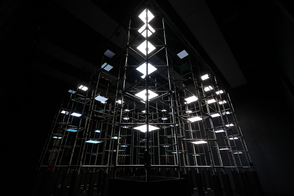 A geometric structure made of scaffolding with illuminated square panels in a dark interior setting.