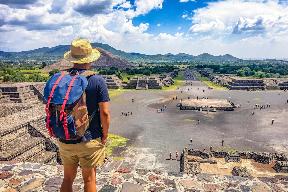 A person with a hat and backpack overlooks the ancient pyramids and ruins of Teotihuacan under a partly cloudy sky.