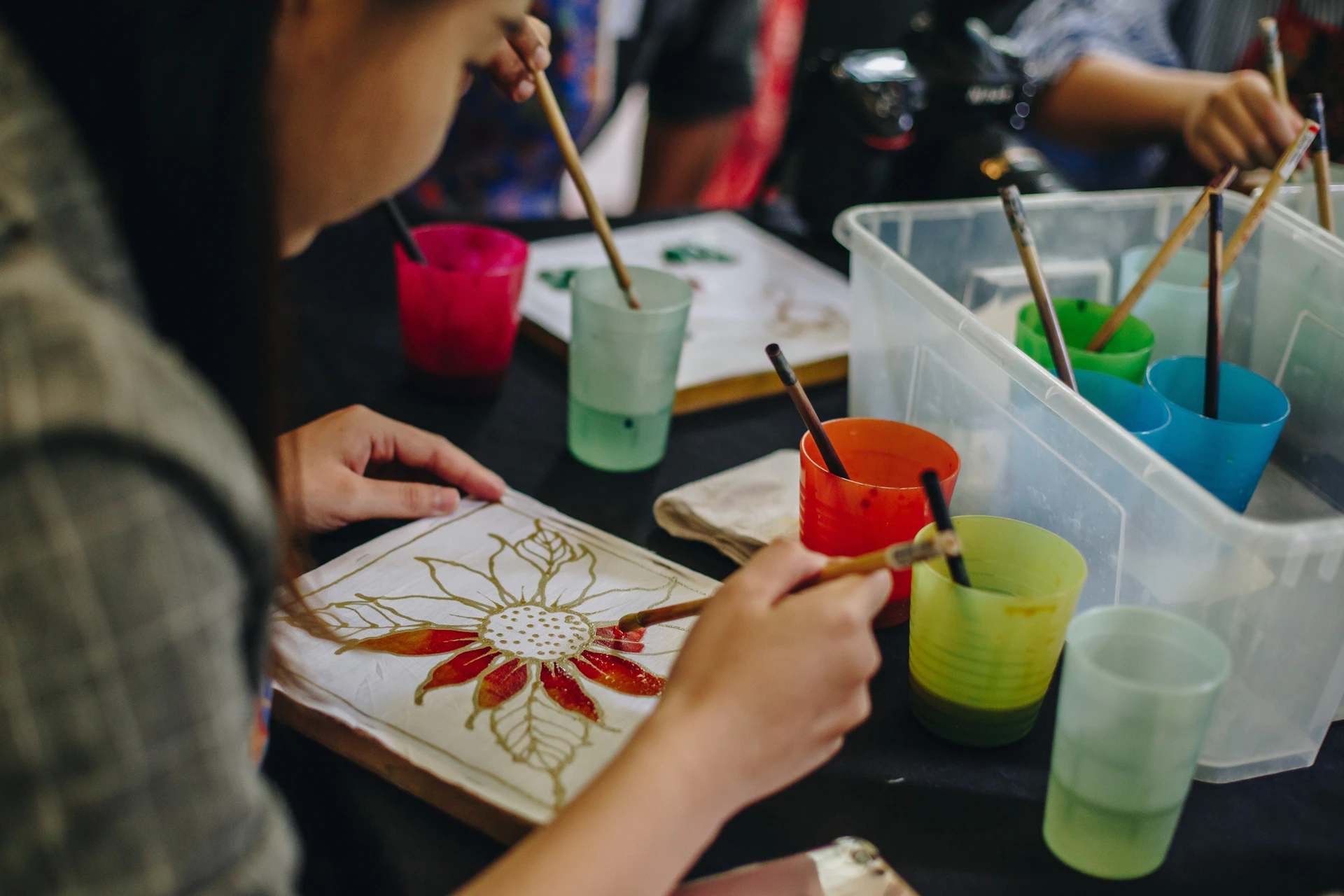 A person painting a floral pattern on fabric, with brushes and colorful cups on a table.
