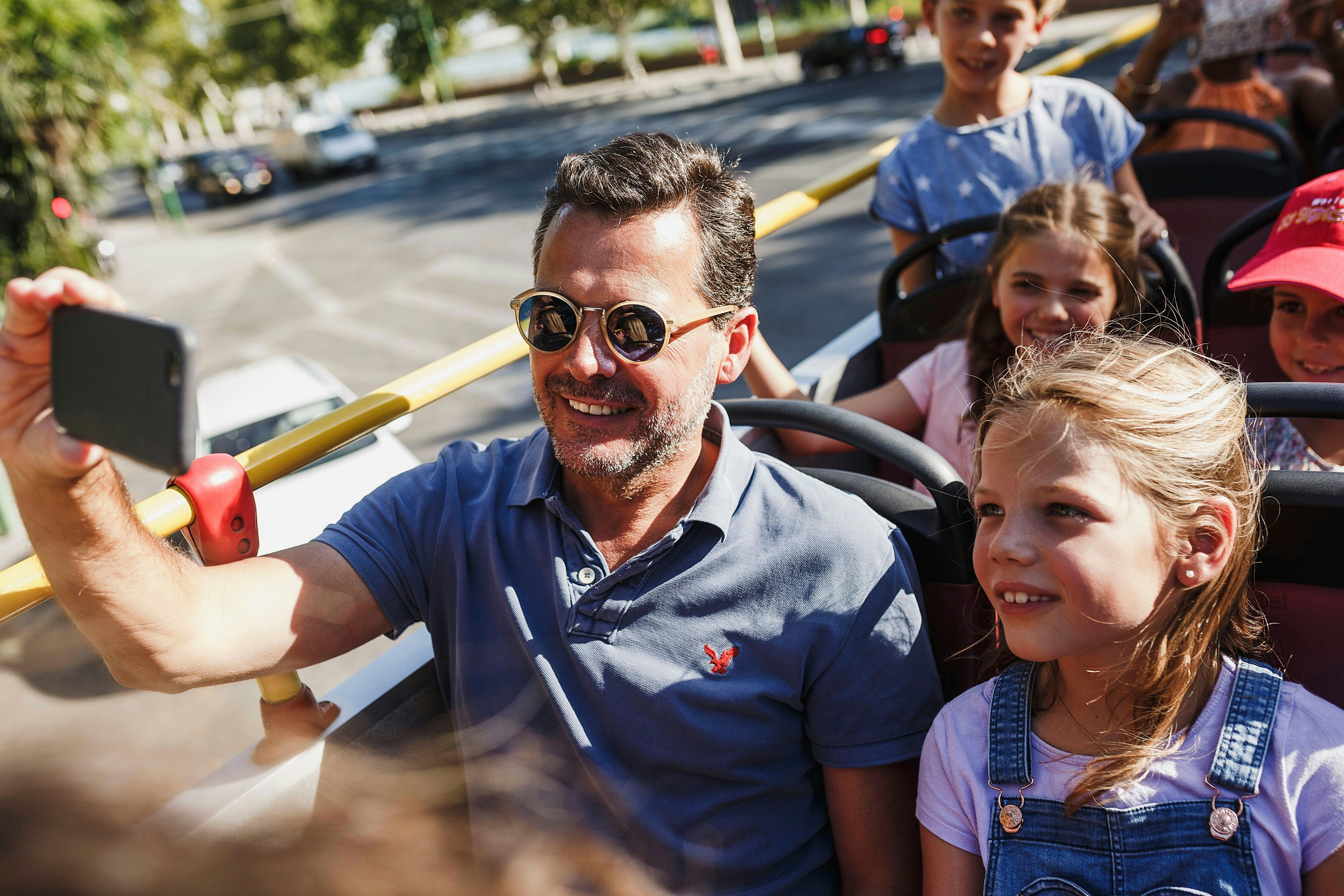 Мужчина и четверо детей улыбаются и сидят в солнечном автобусе с открытым верхом.