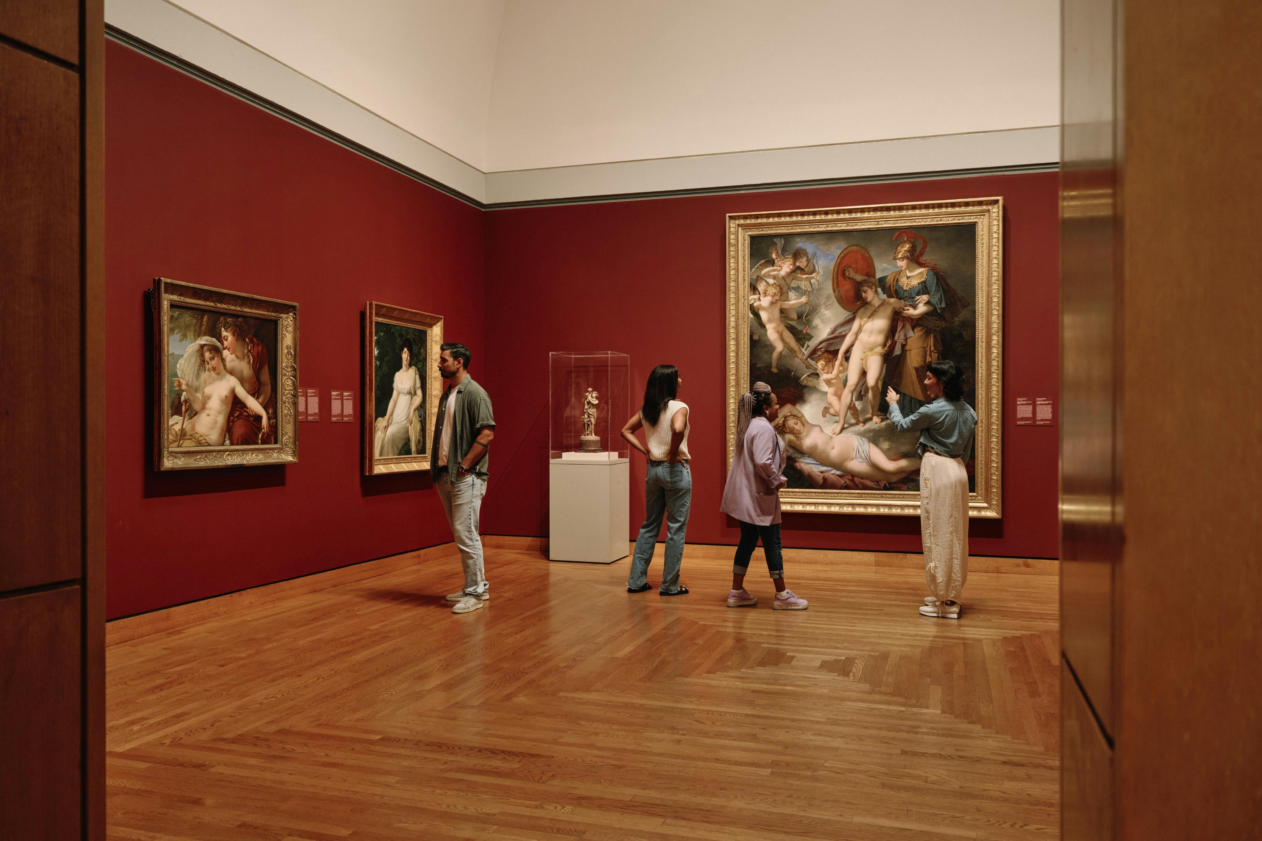 Four people observe classical paintings and a sculpture in a museum gallery with red walls and wooden floors.