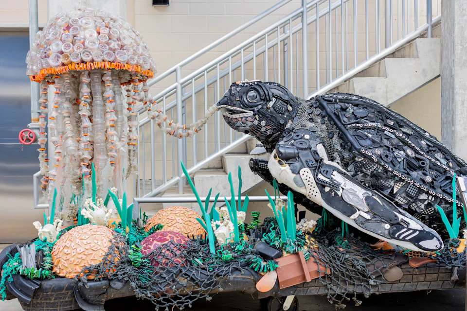 Sculpture of a sea turtle and jellyfish made from recycled materials, set in a colorful coral reef scene with stairs in the background.