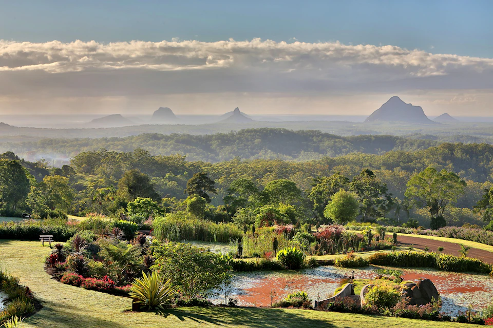 Lush garden with varied greenery, a pond, and distant rolling hills under a partly cloudy sky with mist-covered mountains.