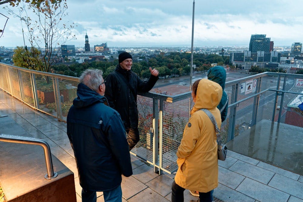 Čtyři lidé v bundách, jeden ve žlutém, stojí na vyhlídkové palubě s výhledem na město. Jedna osoba při mluvení gestikuluje.