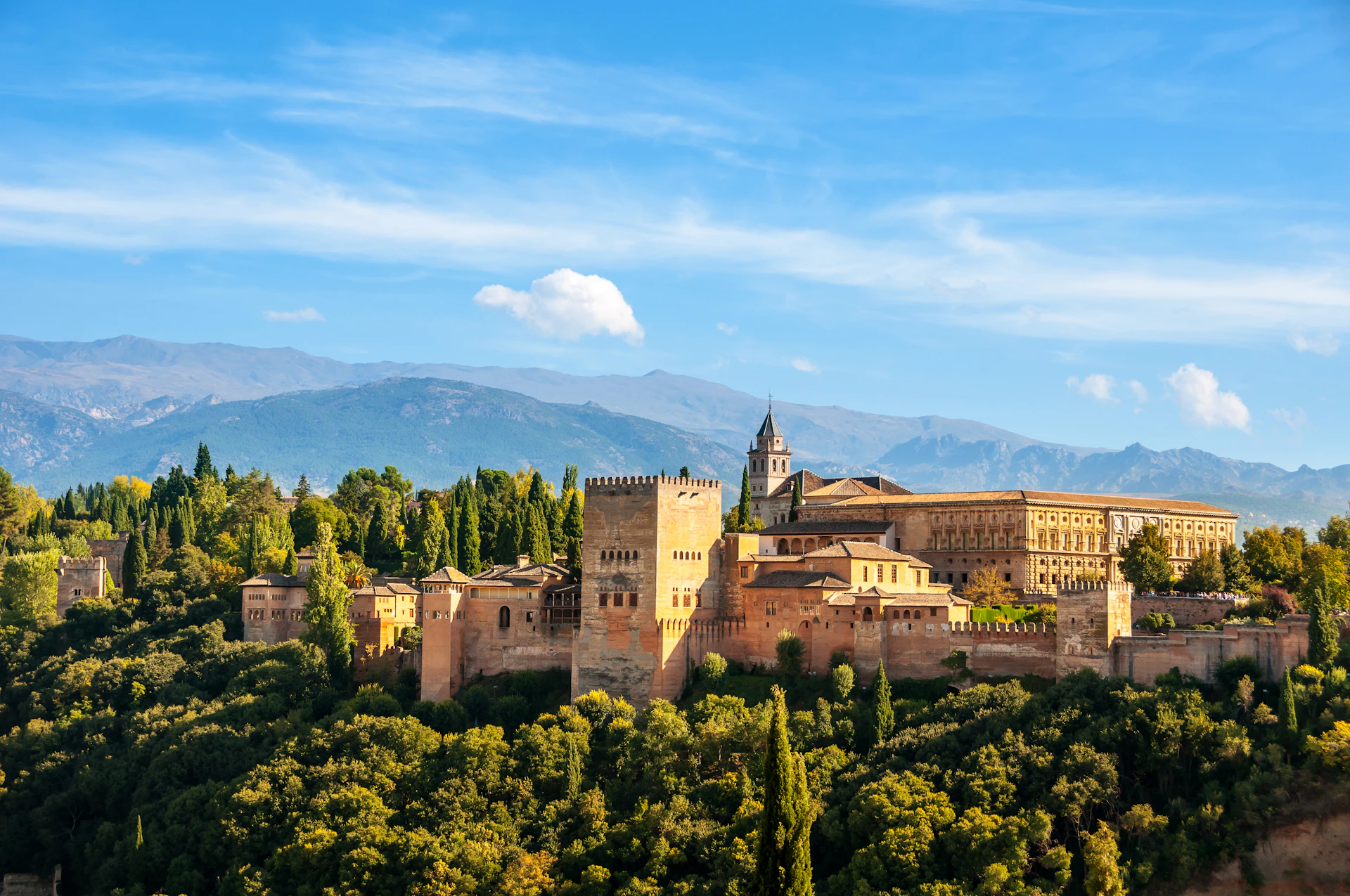 A historic fortress with stone towers and buildings is surrounded by lush greenery, with mountains in the background under a blue sky.