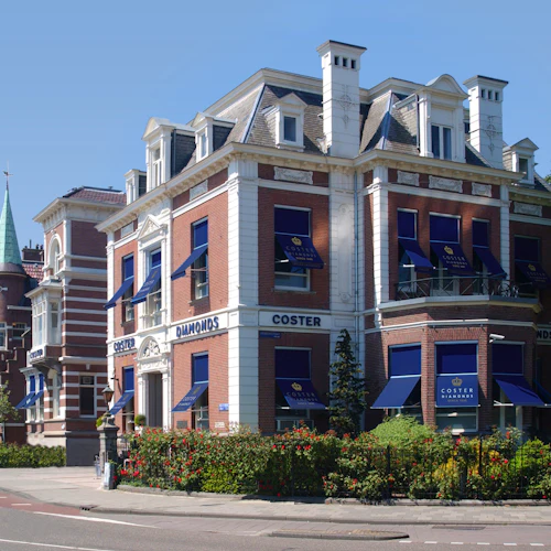 Historisk tegelbyggnad med blå markiser och skyltar med texten "Coster Diamonds" på en stadsgata, med en trädgård framför.