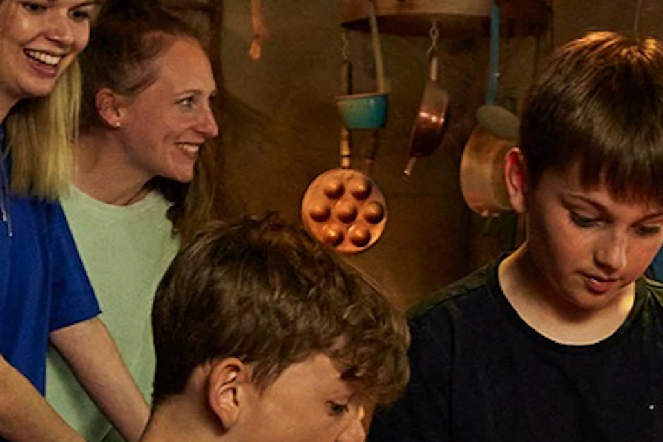 Two children are stirring a large cauldron whilst two adults watch from a dimly lit room decorated with a Halloween theme.