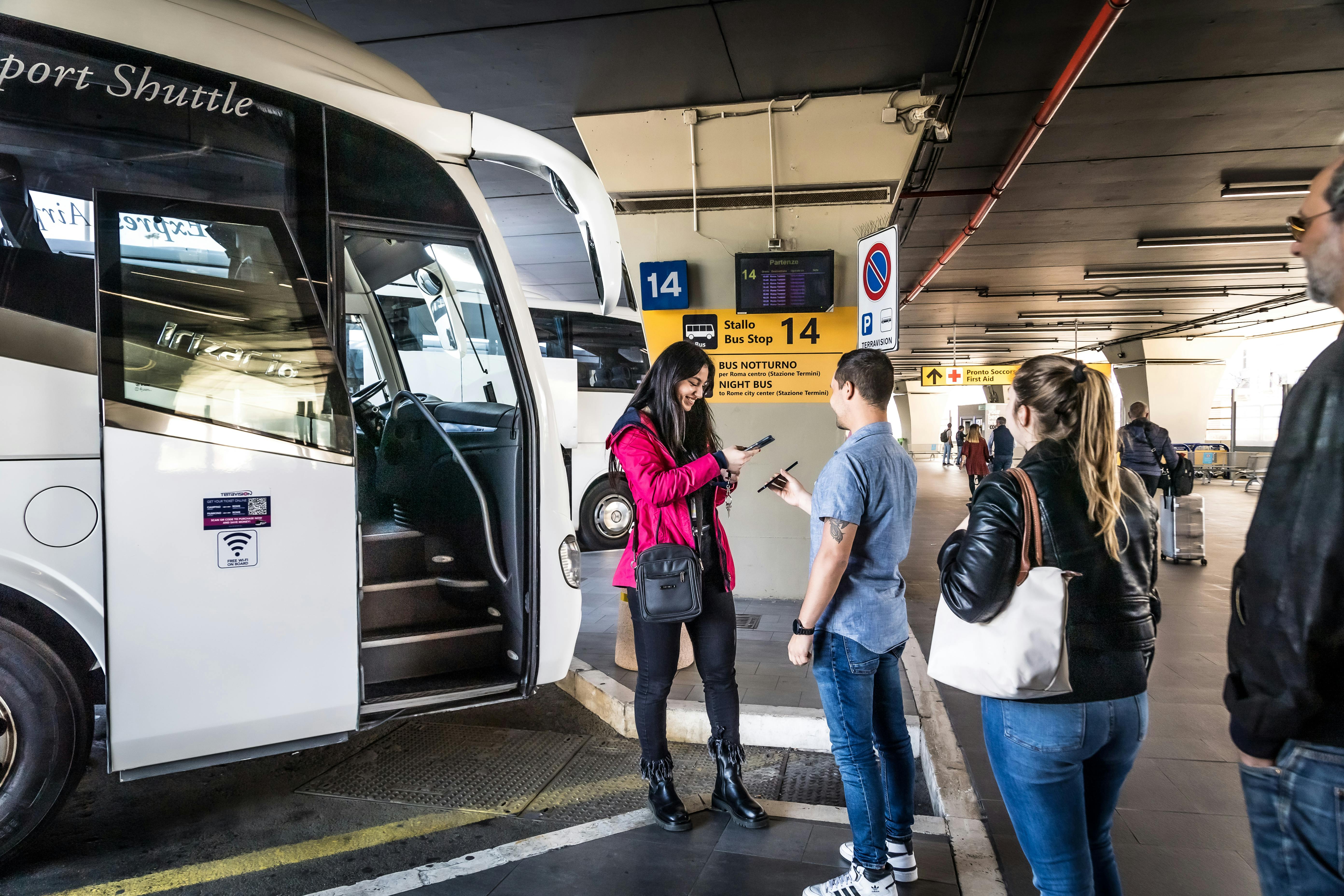 A woman in a pink jacket checks the tickets of two people next to a white bus at a bus terminal marked with signs for stall 14.