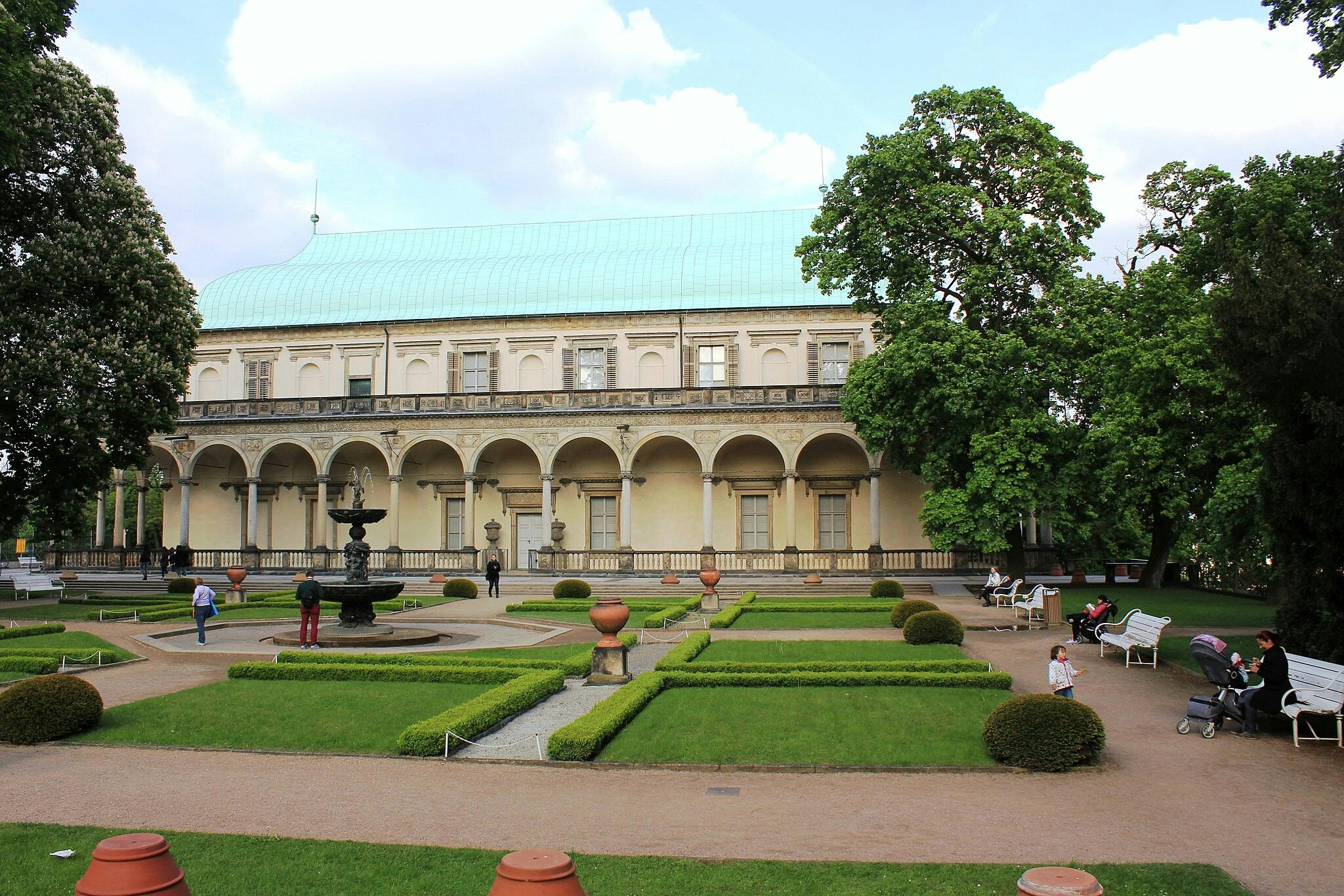 Pessoas a passear e sentadas num jardim com uma fonte em frente a um grande edifício com colunas em arco e um telhado verde.