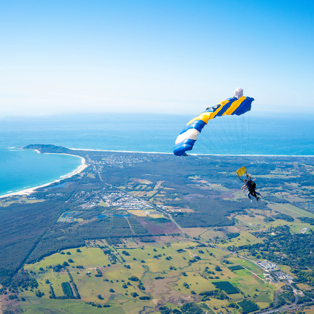 Skydiver descending with a blue and yellow parachute over a coastal landscape with farmland, forests, and ocean in the background.