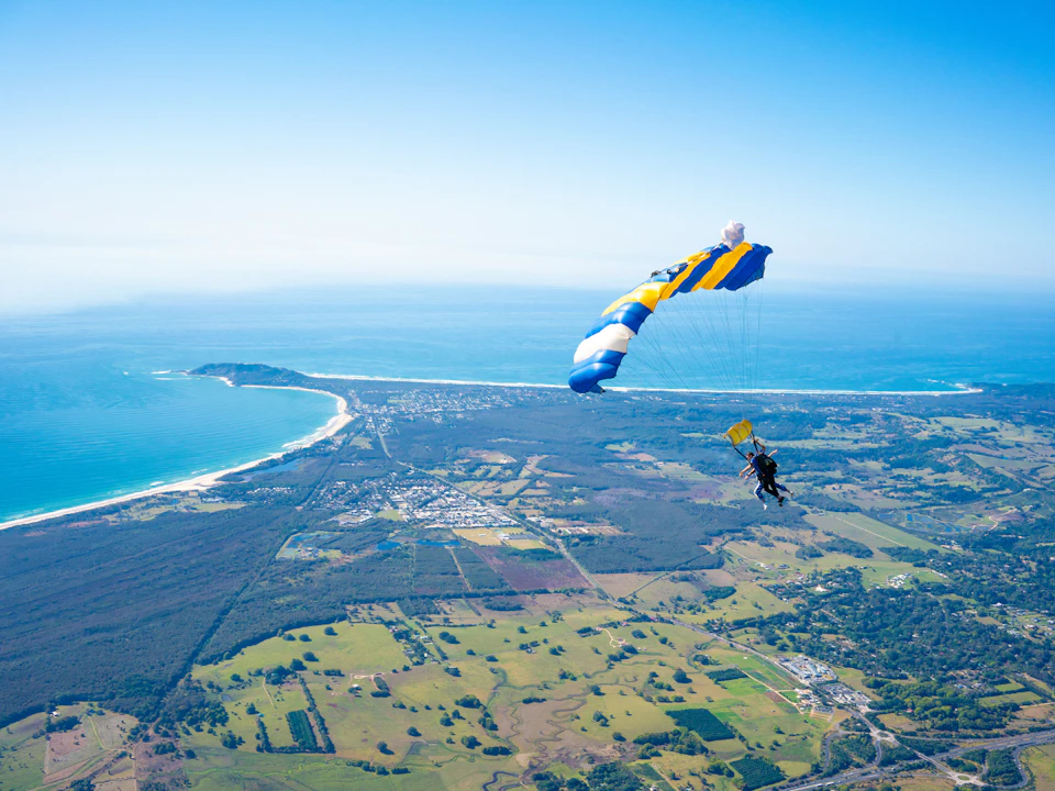Skydiver descending with a blue and yellow parachute over a coastal landscape with farmland, forests, and ocean in the background.