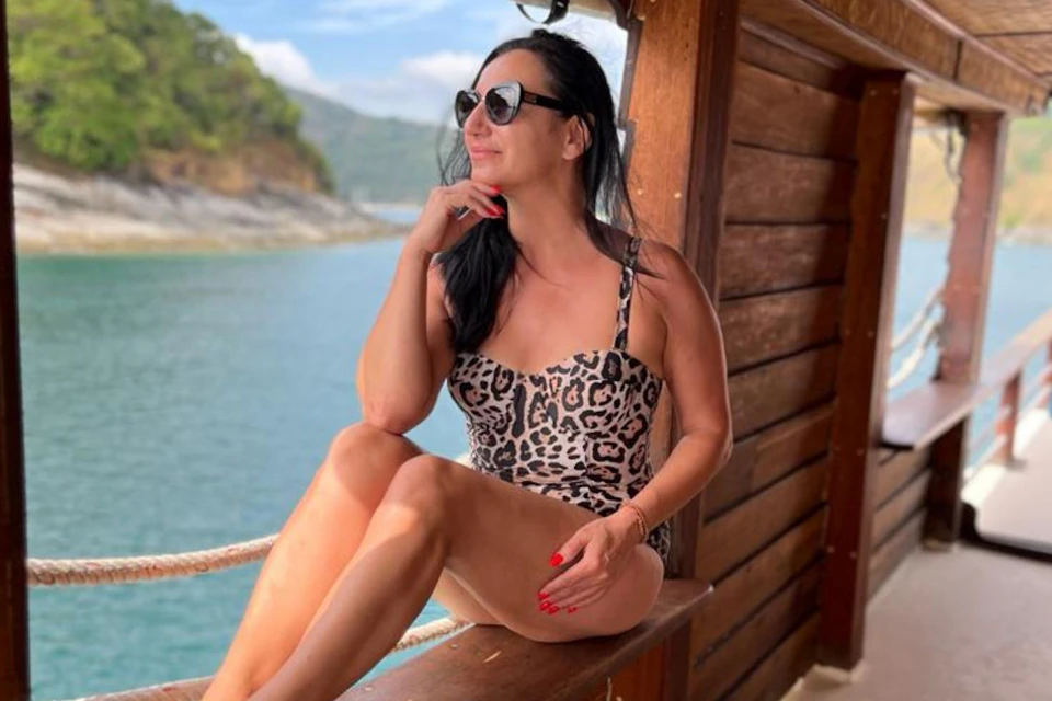 A person in a leopard-print swimsuit sitting on a wooden boat railing near water, wearing sunglasses and smiling.